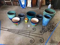 Wide view showing 11 flower pots of different sizes and colors, metal plant holders and trellis on garage floor.