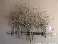 Front view of metal 3D wall art depicting bare trees with branches behind a black fence mounted on a beige wall.