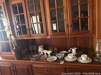 Cut glass bowls, bone china cups and saucers, creamer, sugar bowl, pitcher, two glasses displayed on a wooden cabinet under glass doors.