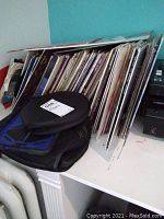 Multiple vinyl LP records stacked upright including various album sleeves and cases packed on a shelf by a teal wall.