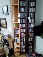 Three tall wooden CD racks filled with jewel cases against a wall alongside empty rack