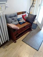 Front view of wooden bench with metal side supports, hinged seat storage and cushions against wall and radiator