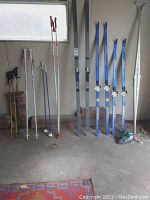 Photo showing five pairs of skis and four pairs of ski poles arranged standing against a wall, including Karhu and Kneissl skis and various poles.