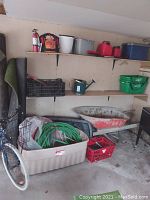 Photo showing various items including red gas cans, fire extinguisher, storage bins on shelves, green crate, watering can, black crate, red wheelbarrow, coiled green garden hoses in plastic bin, and part of a bicycle tire.