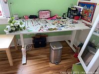 White adjustable height desk with various jewelry and craft items on top, in a green-walled room.