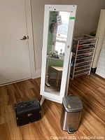 Full view of the white mirrored jewelry cabinet, stainless steel garbage can, and black storage box on a wooden floor.