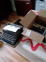 Royal Quite De Luxe C typewriter in its carry case with paper loaded, set next to a cardboard box filled with various books.
