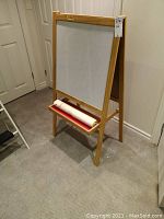 Front view of wooden easel with whiteboard surface and paper roll on tray beneath, placed indoors on carpeted floor near door.