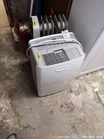 Photo of white portable dehumidifier with control panel on top and handle, placed on basement floor.