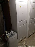 Tall white shelving unit with a closed door next to another similar shelving unit, placed on a concrete floor with a portable heater nearby.