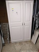 Full front view of the closed white two-door cabinet showing door panels and handles. Cabinet is tall and has a clean look with light scratches visible.