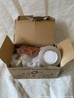 Open box showing one round LED recessed ceiling light with frosted diffuser and silver trim, plus plastic anchors and installation parts inside the box.