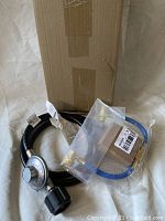 Photo of braided hose with metal fittings and plastic regulator knob, including blue hose with brass connectors in a plastic bag, alongside a cardboard box.