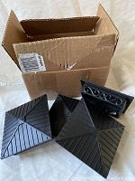 Four black plastic fence post caps with one showing ornate scroll design, placed in front of a damaged cardboard box.