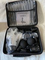 Open carrying case displaying the Neprok massage gun, two large ball attachments, two other types of attachments, and a power charger, all sealed in plastic bags.