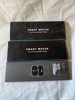 Three black smart watch boxes stacked on fabric background, each labeled as Smart Watch Your Personal Health Tracker.