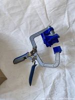 Handheld corner clamp tool with metal frame and blue plastic handles and corners, resting on white cloth background.