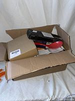Open cardboard box containing various kitchen utensils including a large red plastic spoon and several black and brown boxes.