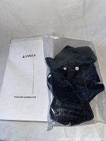 Black Vokka posture corrector in plastic bag, with instruction manual and box behind showing brand and product name.