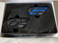 Open box containing two Lexin motorcycle Bluetooth intercom communication devices, one with blue accents and both with multiple buttons labeled FM and others.