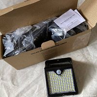 Open box showing six black solar motion sensor LED lights individually wrapped in plastic packaging with an instruction manual included.