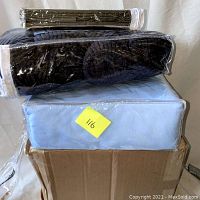 Stacked items in plastic packaging: dark pillow holder on top, light blue boxed chair covers below, in cardboard box.
