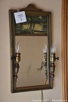 Front view of antique lighted mirror showing ornate frame, scenic painted panel, mirror with black spots, and two candle-style light fixtures.