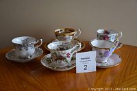 Five Royal Albert teacups with matching saucers and a small plate on a wooden table; includes assorted floral designs and one Lady Hamilton set with rich brown and gold pattern.