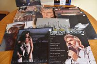 Photo showing multiple vinyl LPs stacked and overlapping on a wood table, featuring Kenny Rogers Greatest Hits and other artists.