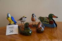 Six bird figurines on wooden surface with MaxSold lot number sign, includes ceramic ducks, bird figurines on stands, plastic duck napkin holder.