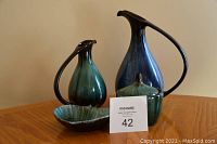 Four pottery pieces including leaf dish, sugar bowl with lid, small green jug, and large blue jug displayed on a wooden table.
