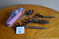 Five pairs of metal scissors in varying sizes and styles displayed with a painted wooden scissor holder on a wooden table.