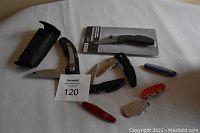 Eight assorted pocket tools including knives and multi-tools laid out on white surface with MaxSold lot card visible, one tool in original packaging, one with sheath.