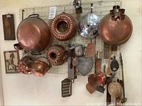 Overview of wall-mounted copper pots, molds, and kitchen utensils arrayed on a metal grid.