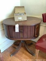 Front and top view of the oval wooden side table showing the sliding tambour doors with an old clock face design.
