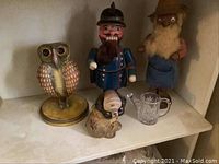 Shelf with brass and copper owl statue, wooden nutcracker soldier figure, wooden gnome figure, cast iron pug coin bank, and small glass cup.
