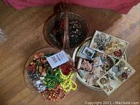 Three baskets/trays showing assorted necklaces, beads, brooches and earrings across a wooden floor