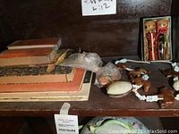 Shelf with several vintage Asian art paper booklets in orange and beige covers, and assorted costume jewelry items including carved wooden camel beads, plastic beads, a shell pendant, and a small figurine in plastic bag.