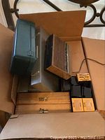 Photo showing a brown cardboard box filled with assorted film slide containers, plastic slide trays with yellow labels, two wooden boxes, a green metal container, and a vintage alarm clock with a wood grain front and metal sides.