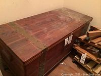 Front and side view of rectangular cedar chest with metal strip reinforcements and visible wear including scratches. Numbers are noted on chest tags attached to the side.