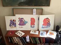 Photo of larger signed Barbara Lavallee Indigenous American art prints on boards showing figures of women and children in traditional dress, with visible staining on some prints.