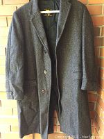 Full front view of the wool tweed coat hanging against a brick wall, showing button closure, lapels, and flap pockets.