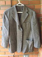 Front view of a gray checked wool tweed jacket hanging on white hanger against brick wall.