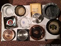 Top-down photo showing 12 ashtrays in various materials including glass, metal, wood-tone, and marble-style, showcasing different emblems and designs