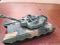 Green camouflaged remote control tank with turret, cannon, and a red antenna. Displayed on a brown surface.