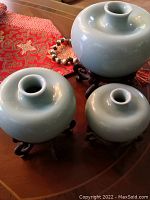 Set of three green-glazed porcelain vases of varying size, each with wooden carved stands, placed on a wooden table with a red decorative cloth nearby.
