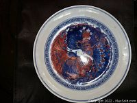 Top view of decorative porcelain plate with butterfly and floral design in blue and reddish-orange colors, bordered by Greek key pattern.