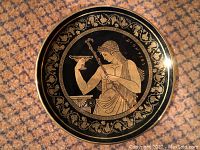 Close-up of the metal plate featuring a seated Greek figure holding a cup in gold and black