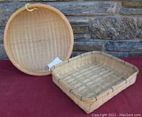 Photo of two bamboo baskets, one round and one rectangular, placed on a red textile backdrop against a stone wall. The round basket has a continuous weave pattern.