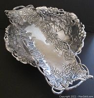 Two Arthur Court aluminum serving pieces featuring grape and leaf motifs in relief: one rectangular tray measuring 19 inches by 6 inches and one rounded bowl measuring 14 inches wide by 4 inches high. Both pieces show detailed raised grapevine designs and surface wear consistent with use.